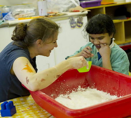 a female teacher and a young autistic
                  boy play with a red container filled with white flour