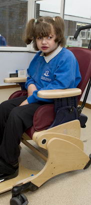 A young girl sits in a supportive
                  chair whilst her female teacher straps down her feet