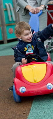 A boy drives a toy car around a playground,
                  pushed by an adult