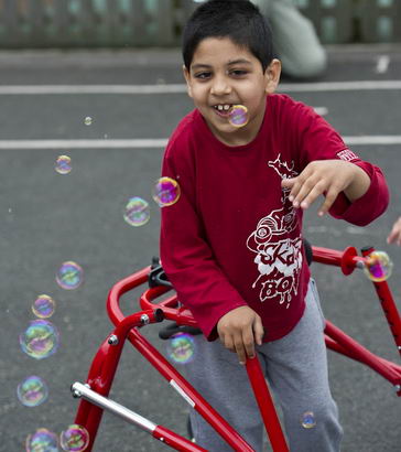 A boy in a playground with walking
                  support smiling at airborne bubbles