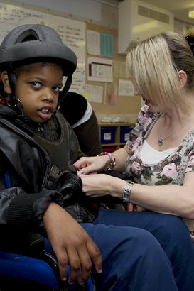 A young black boy with protective
                  headgear in a wheelchair is helped by a teacher