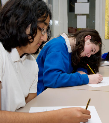 A young girls takes a test alongside
                  her teacher using a personal computer