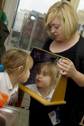 A young girl looks at herself
                  in a mirror held by a member of staff