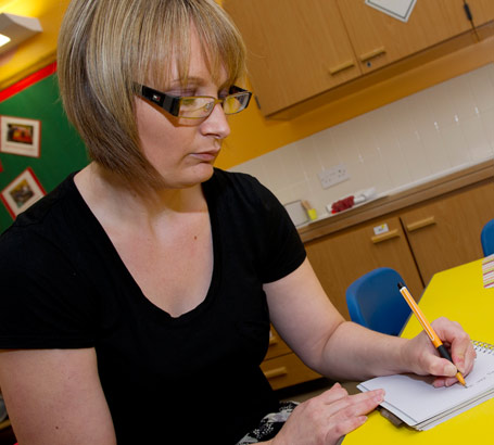 a teacher wearing glasses sits
                  at a table taking notes on one of her pupils