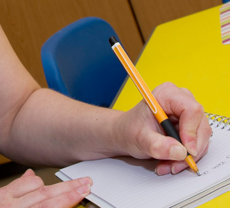 a teacher wearing glasses sits
                  at a table taking notes on one of her pupils