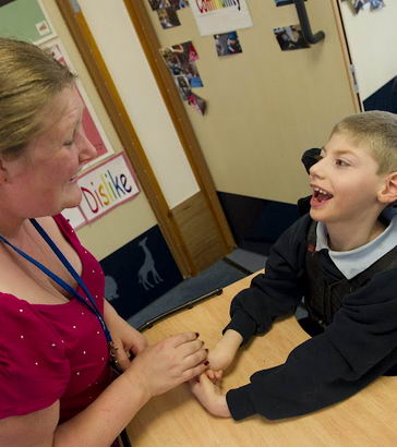 A boy in a specialist chair smiling
                  with his teacher