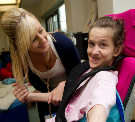teacher holds the arm of a girl in a specialist
                  chair smiling at the camera
