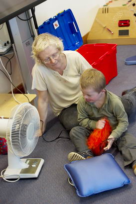 A teacher and a young boy seated
                  on the floor with a cushion and a fan