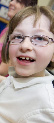 young
                  girl in white top smiles at camera, sitting next to assistive device