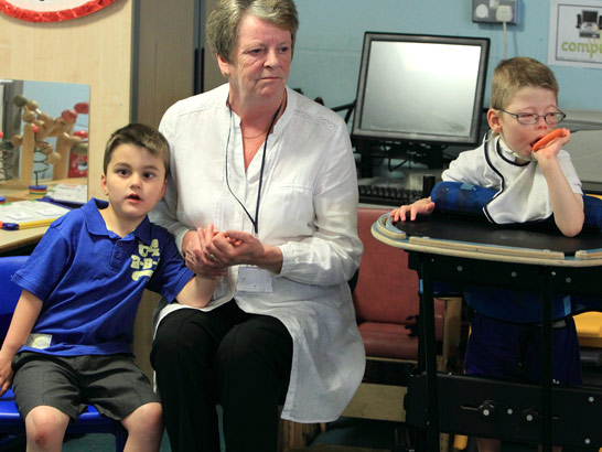 An older female teacher comforts, supports
                  and holds the hand of a young boy width=