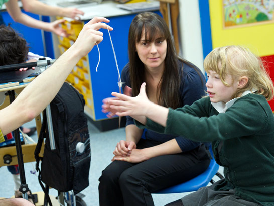 a young girl spins a pendulum
                  whilst her teacher and classmates look on