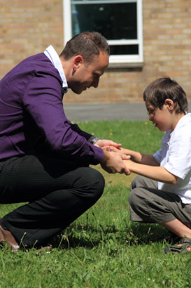 A teacher holds a young boy's hands in
                  the playing fields behind their school
