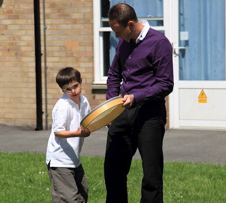 A boy and his teacher stand on the grass
                  with a drum