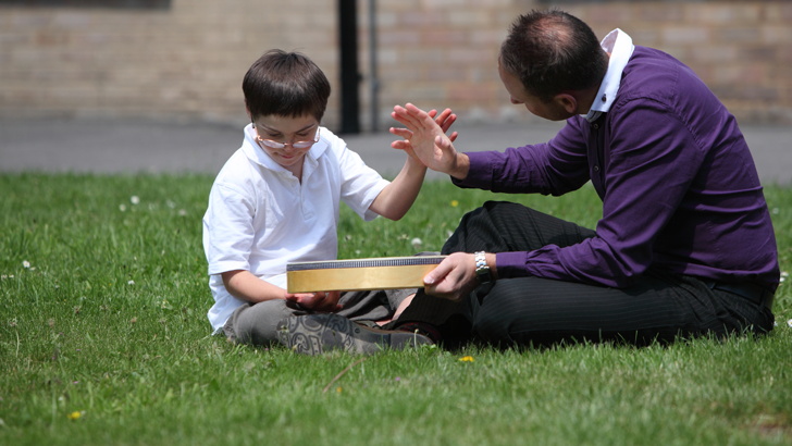 A boy and a therapist sit on
                  the grass with a large drum