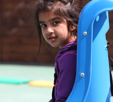 A girl sits in a blue seat in a playground,
                  smiling at the camera