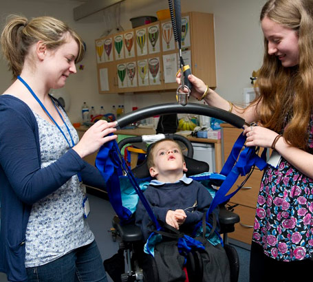 Two carers lower a boy into a wheelchair
                  using a hoist