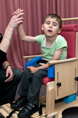 A teacher holds the hand of a boy
                  in a specialist chair