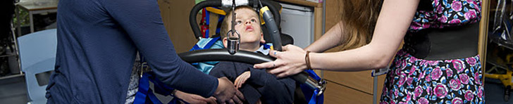 A boy being lifted out of a wheelchair
                  with a hoist