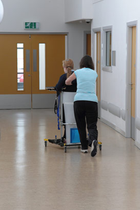 A girl in a mobile standing frame being
                  moved by a carer