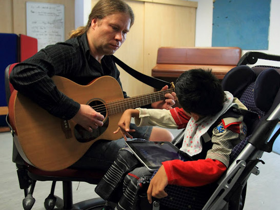 Disabled boy listening to a carer playing
                  the guitar.