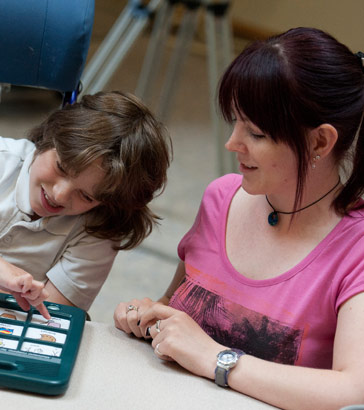 A teacher watches a boy select an image on a communication device