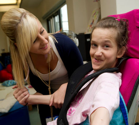 teacher holds the arm of a girl in a specialist chair smiling at the camera