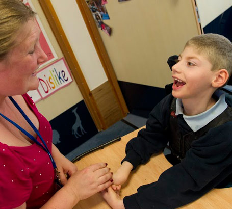 A boy in a specialist chair smiles at his teacher]